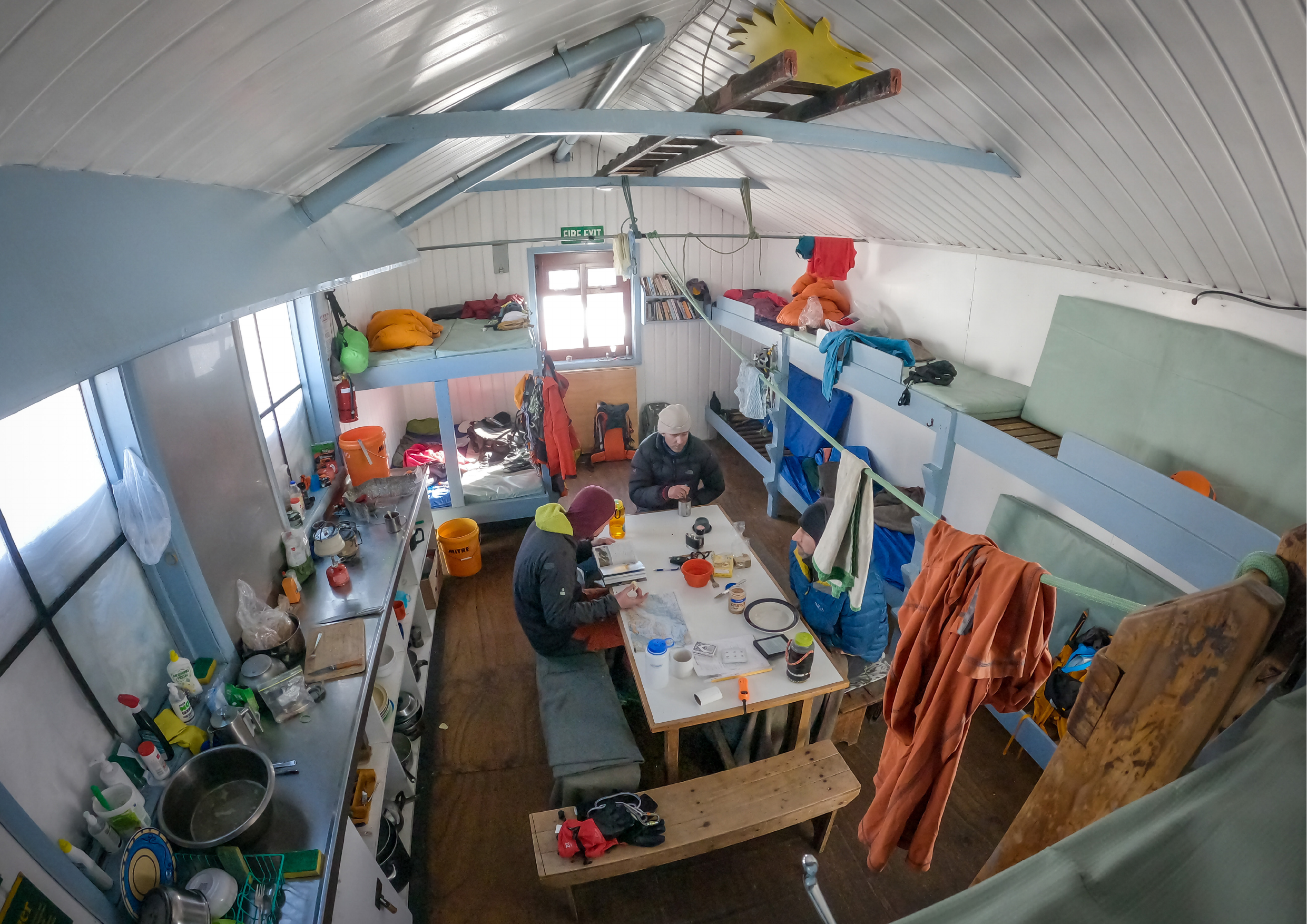 Inside Kelman Hut — two team members at the table, gear spread through the cosy mountain hut