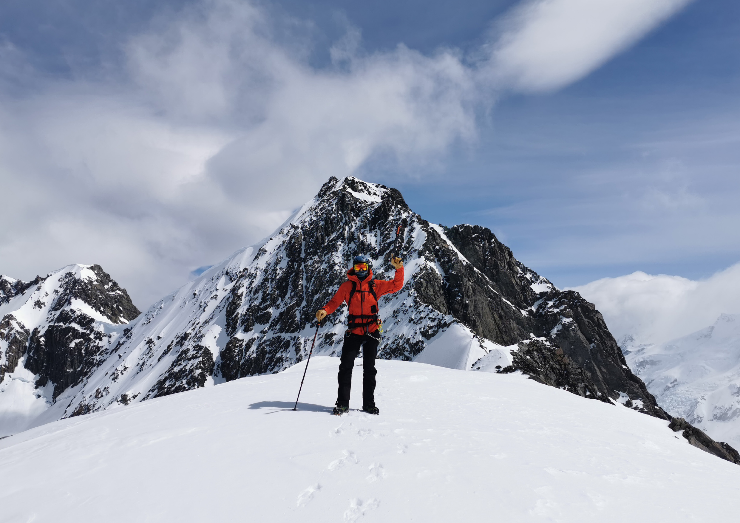 Climber on a summit with a rocky peak rising behind, surveying the Southern Alps